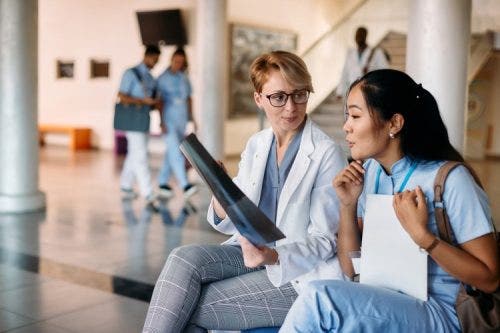 A nurse preceptor sits on a hospital bench discussing an x-ray with a new nurse.