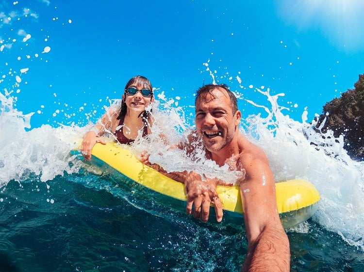 A father and daughter on a yellow-and-blue raft are riding the ocean waves at the beach.