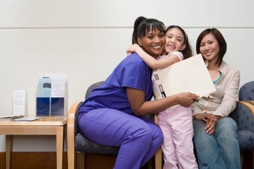 A nurse holding a clipboard hugs a child while another person smiles and looks on.